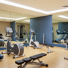 A well-lit, modern gym with various exercise equipment. The room has a row of treadmills on the left, a set of dumbbells in the center, and weight machines on the right.